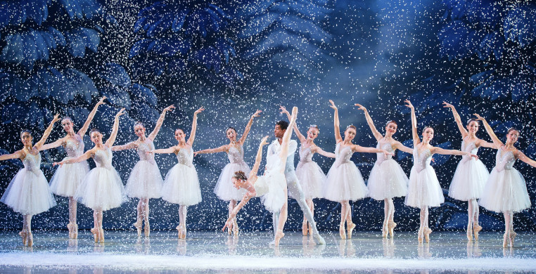 A magical ballet scene from "The Nutcracker" featuring the Snow King and Snow Queen at center stage, surrounded by graceful ballerinas on pointe. Delicate snowflakes fall gently around them, creating a shimmering winter wonderland. The dancers' flowing co