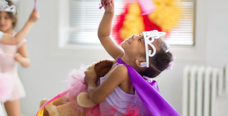 A young child in a creative movement class, wearing a purple leotard, red tutu and tiara, is gracefully dancing in the bright open studio. The child is mid-movement, arms extended, with a look of joy on their face.