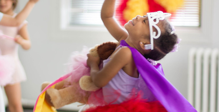 A young child in a creative movement class, wearing a purple leotard, red tutu and tiara, is gracefully dancing in the bright open studio. The child is mid-movement, arms extended, with a look of joy on their face.