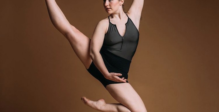 Dancer in olive green leotard and black shorts jumping with right leg extended upward.