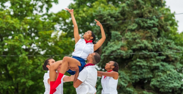 "Journey" Pasteur Park 2021. 3 dancers in white collared shirts, blue skirts or pants, red ties or neck scarves, and red socks stand in front of green trees lifting a lady dancer to their shoulders. She is pointing both hands over her head to the right.
