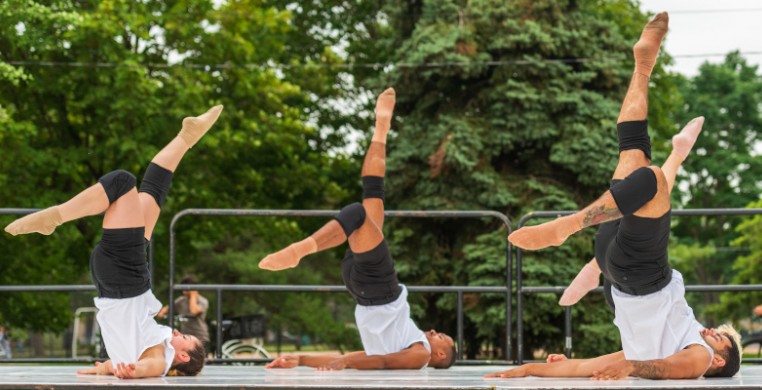 "My Apologies, Mozart" at Pasteur Park 2021. 3 dancers wearing white, sleeveless tux shirts, black shorts, and black knee pads are on an outdoor stage in front of green trees. They are on the floor, kicking their legs over their heads in a shoulder stand.