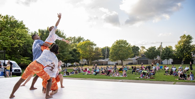 5 dancers on an outdoor stage. They are wearing orange, blue, and white costumes and supporting a small white woman dancer as she reaches to the sky