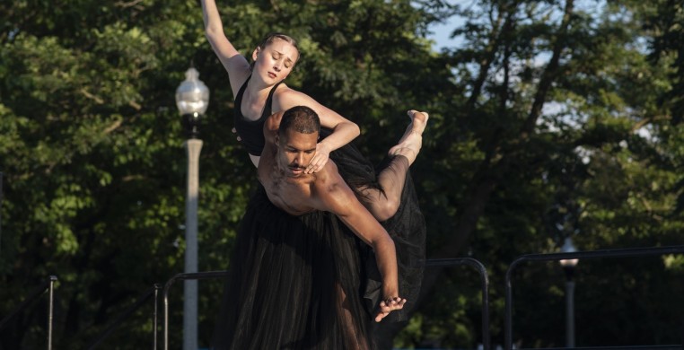 Two dancers wearing black tulle skirts are in a lift. Sam, a tall black male with a mustache, is lifting Tori, a tall white woman with blonde hair, on his back. Sam is looking down while Tori and reaching and looking up