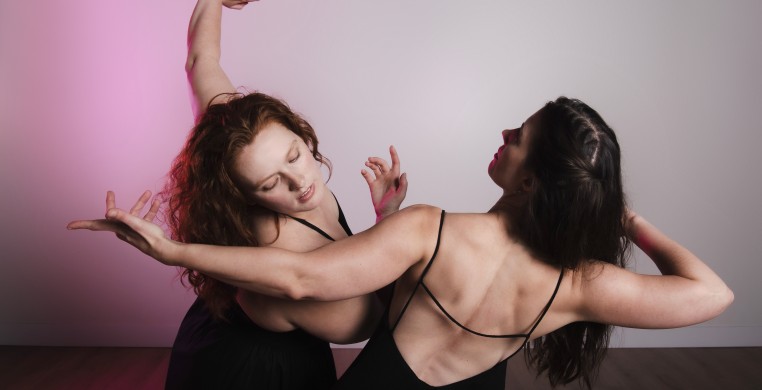 Two dancers in black fabric - one reaching across the space with the other hugging close behind