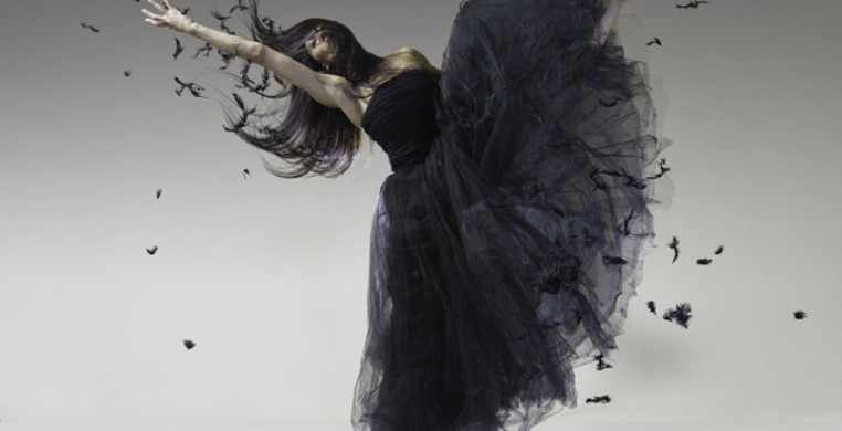 woman in a black long tutu and black long hair throwing feathers in the air while kicking one leg up