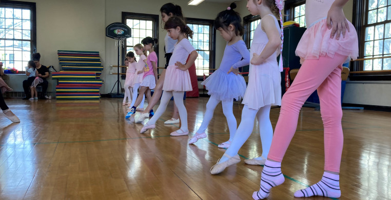 Youth dancers in various dance outfits stand in a line with one foot in front tendu. The dancers are in a busy dance studio with wood floors.