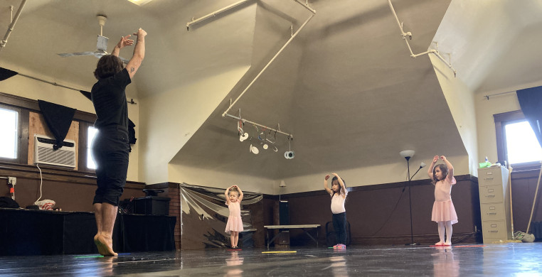 A white trans-masculine person in black clothes demonstrates releve for three youth dancers in various ballet outfits in a dance studio with black floors and wood walls. Everyone is holding their arms overhead in ballet fifth position.