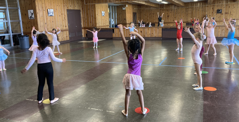 A class of youth dancers in various dance outfits stand in a circle waving their hands in the air in a large room with green tile flooring and mirrors on the wall.