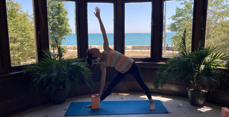 A person in fitness clothing poses with a blue yoga block and blue yoga mat on tile flooring. A large window and view of the lake can be seen behind them.