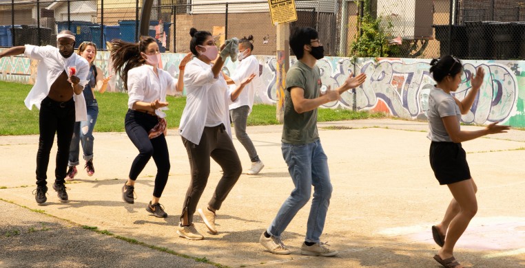 Image of seven people dancing in a line outside on a basketball court with a mural in the background