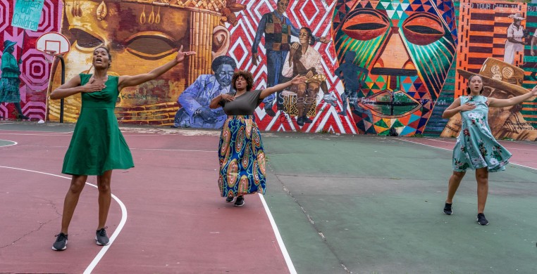 Three dancers in front of a large mural wearing colorful dresses, standing on a basketball court with arms outstretched