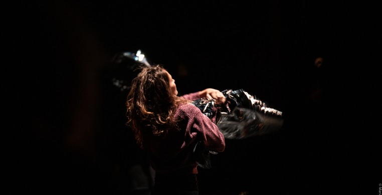 A woman with long brown hair and a dull pink knit sweater, seen from behind and lit from above, hugs a large sheet of silver reflective mylar in a pitch black room. 