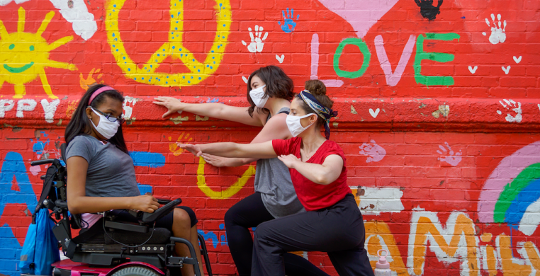 three dancers wearing white masks against a mural on a brick wall. One dancer in a power chair is on the left and two dancers lunge on the right with arms outstretched. Photo Credit: Justin Cooper 
