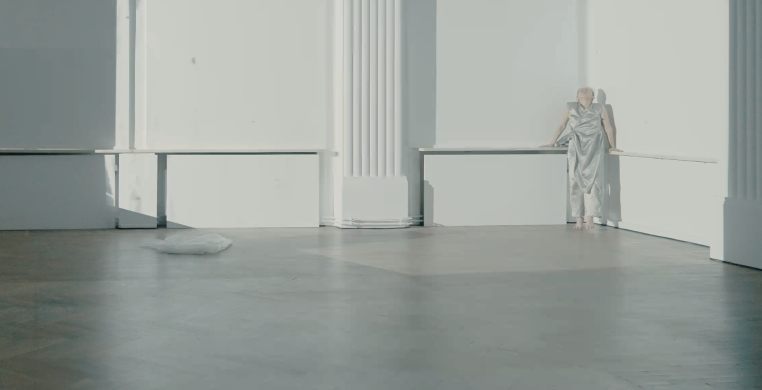 A dancer in a silver costume at the corner of the large room in white projection light.