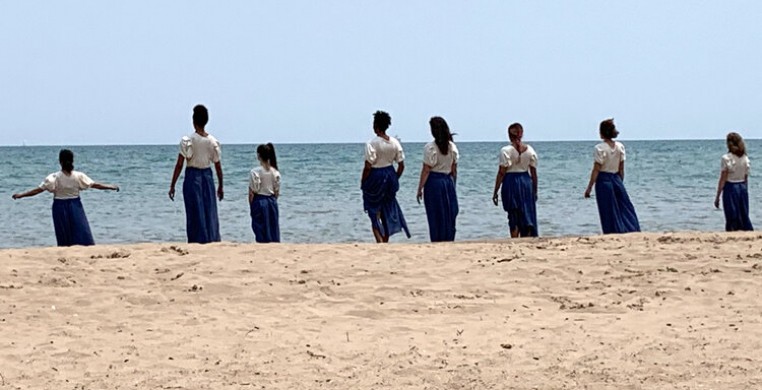 8 dancers in blue and white tunics walk from the beach into the water, against the backdrop of a blue sky. 