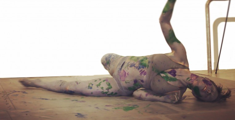 Woman painted all grey with splotches of colored paint dances on floor with arm extended