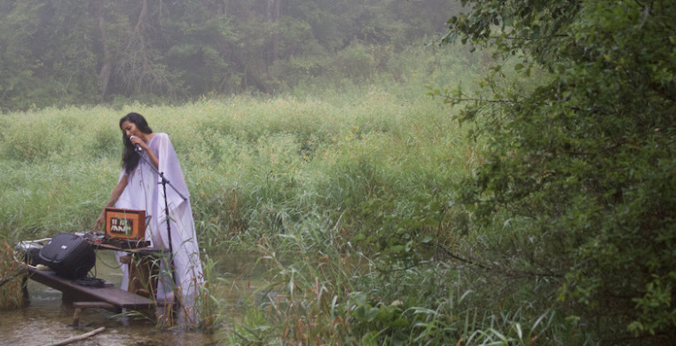 A woman performs into a microphone amid wetlands.