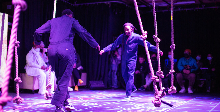 Two people facing each other among a purple-lit space full of thick, knotted ropes hanging down.