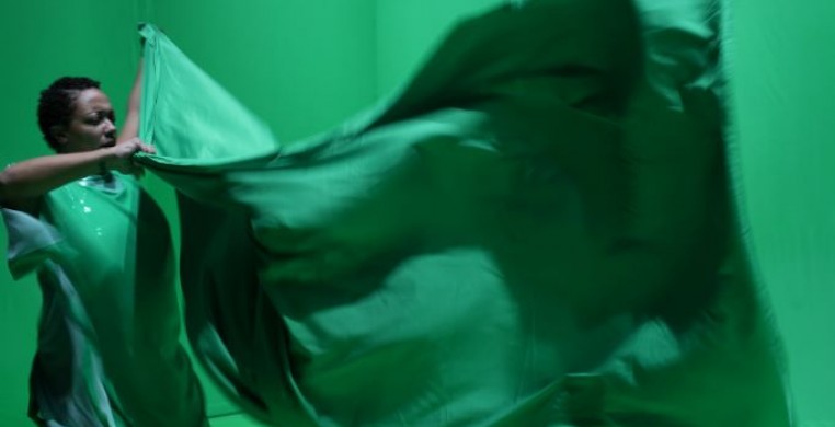 Still image of Black performer with curly hair in green costume holding the corners of a piece of green fabric.