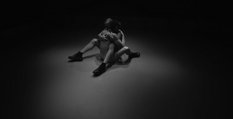 Two performers, facing each other and sitting on the floor, embrace by holding each other in their arms.