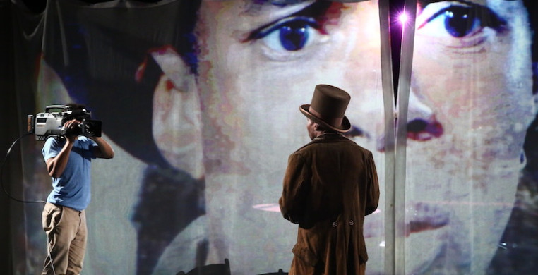 A person turned away from the camera in a top hat looks towards another person holding a video camera and a large screen displaying a man's head