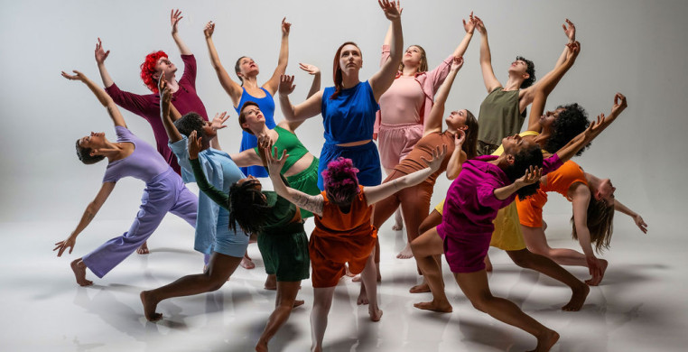 Dancers in multicolor costumes stand in a circle and bloom outward, while a single dancer in the center reaches toward the sky.