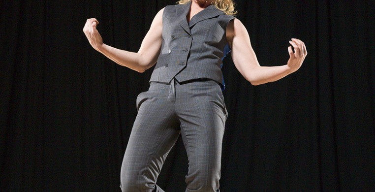 Sara Zalek in the center of a black background, on a wood floor, wearing a matching set grey/blue vest and pants. Their knees are bent in a plie, their weight on their left, right foot just behind. Their arms out to the side with elbows relaxed, palms up.