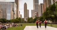 "No More Games" at Lakeshore Park 2018. Three lady dancers in maroon tank tops and black pants stand on a white outdoor stage in the middle of a green field surrounded by tall city buildings. There is an audience in the grass watching the dancers.