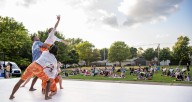 5 dancers on an outdoor stage. They are wearing orange, blue, and white costumes and supporting a small white woman dancer as she reaches to the sky