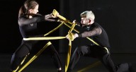 Two dancers in black face each other while intertwined with yellow fabric ribbons