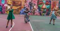 Three dancers in front of a large mural wearing colorful dresses, standing on a basketball court with arms outstretched