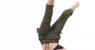 A white trans-masculine dancer with long brown hair and facial hair in a black crop top and army green pants balances on one shoulder with legs extended overhead in front of a white background.